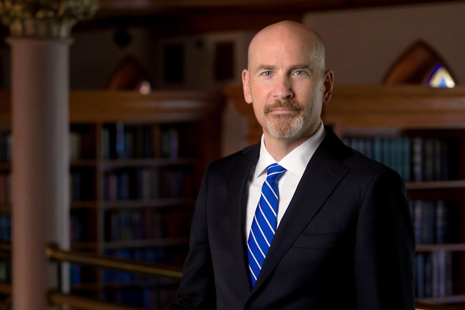 A well-dressed bald man in a suit and tie stands before a collection of bookshelves, showcasing a variety of books.