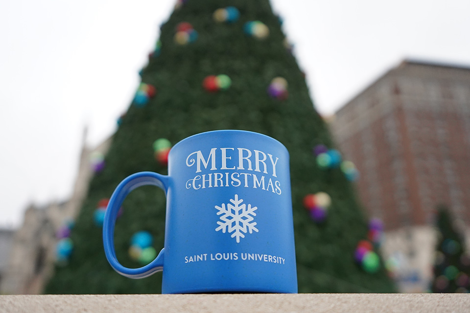 COTQ mug A mug says Merry Christmas Saint Louis University with an image of a snowflake on it as it rests in front of a large Christmas tree outside.