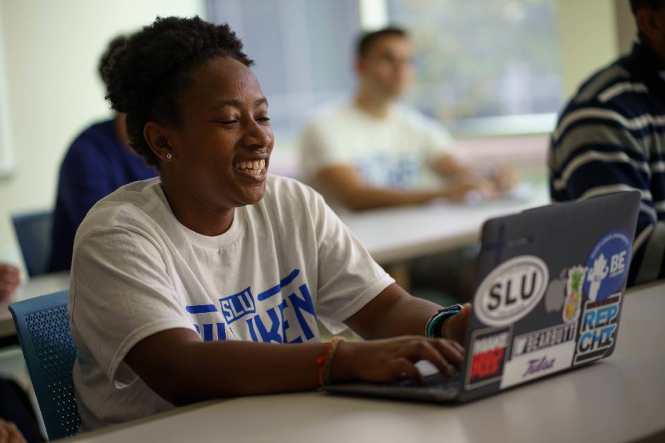 A student wearing a Billiken t-shirt sits in a classroom, smiling while typing on a laptop.