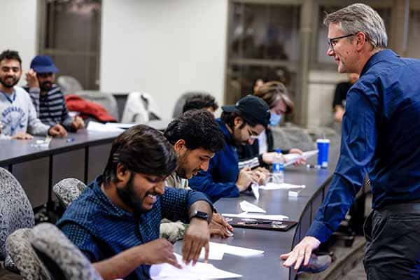 Students sit and work on an assignment  in a lecture-hall style classroom. A male faculty member stands in front of them.