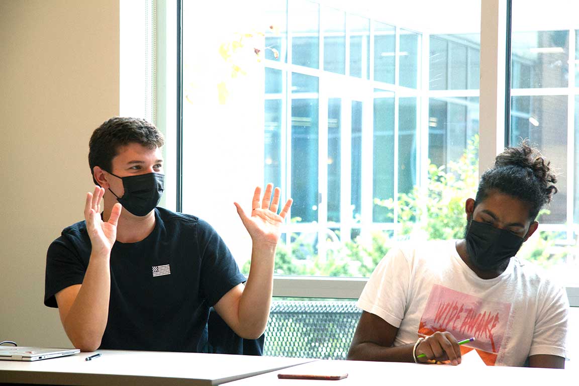 Two students in masks sit at a table in a classroom, one with both hands raised palm side up. 