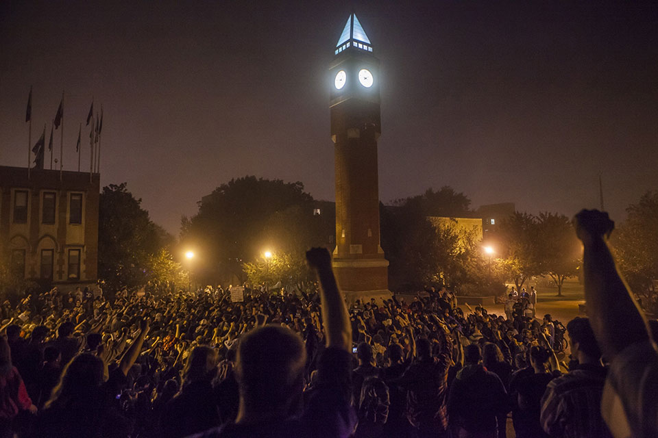 The SLU clock tower is lit up at night while surrounded by hundreds of students with fists raised in the air.