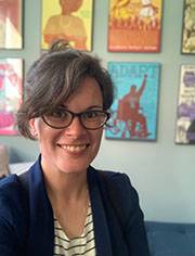 Allison Brewer, headshot. They are wearing a navy jacket over a blue and white striped top and glasses and smiling at the camera.