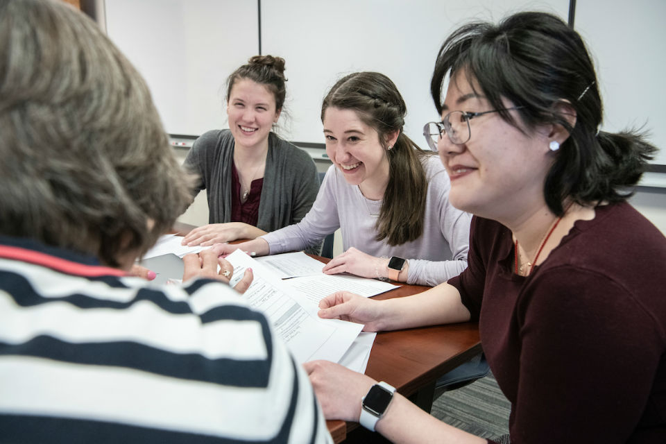 Four students sit around a table in a classroom talking and smiling.