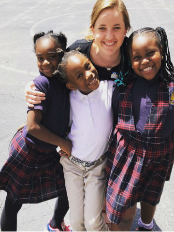 A B T C member stands on a concrete playground with her arms around the shoulders of three students wearing school uniforms.