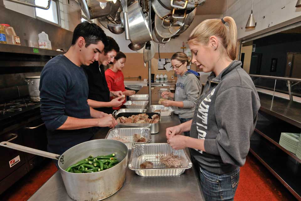 Students standing at a counter in the middle of a  a commercial kitchen, preparing food.