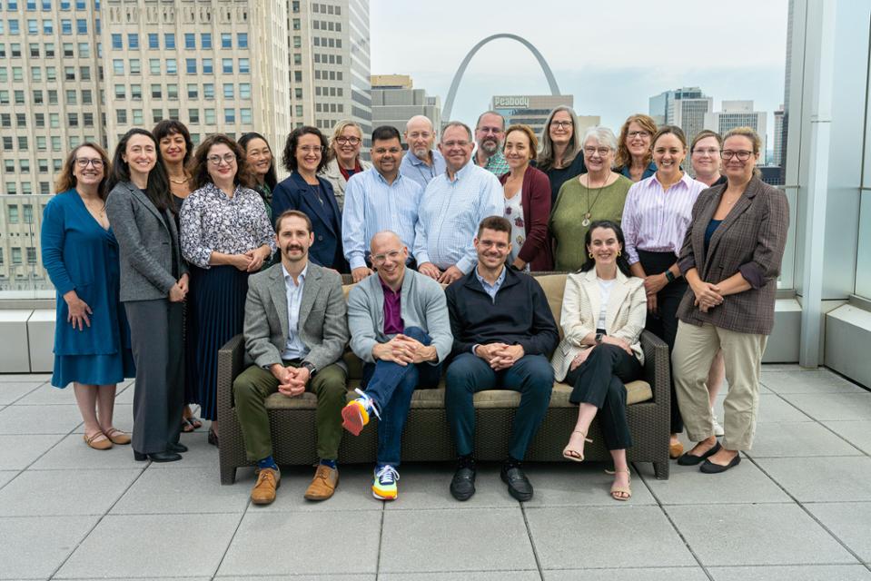 A group of people pose for a photo on a rooftop patio with the St. Louis Arch in the background