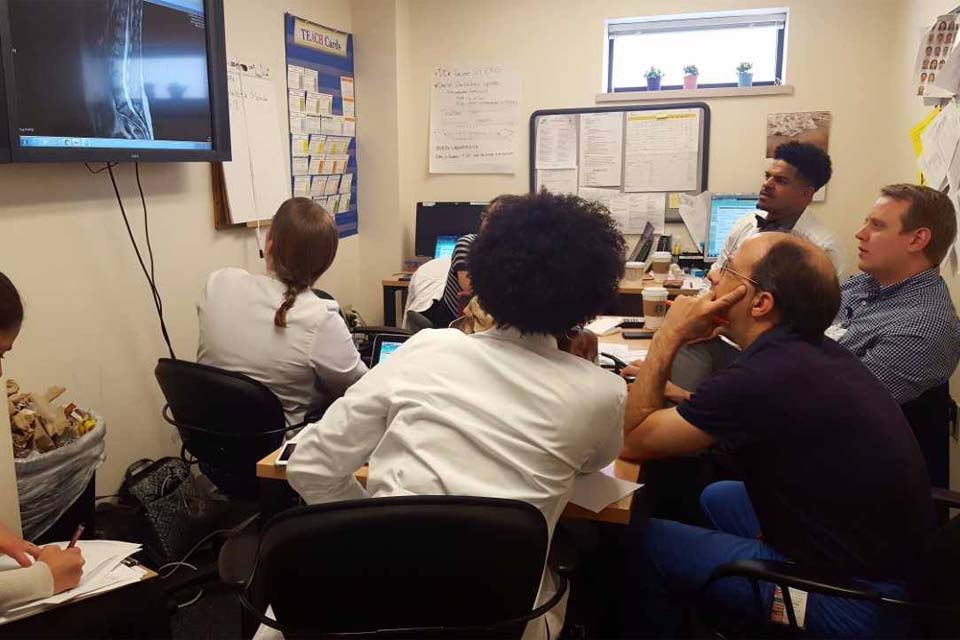 a group of doctors sitting around a table looking up at a tv 