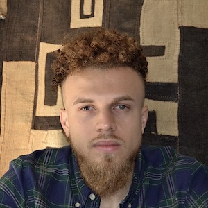 A shoulder-length photograph of an adult man with auburn hair in tight curls, close cropped along the sides, a moustache and a well-trimmed, triangle-shaped beard, gazes calmly straight ahead. He wears a blue and green flannel shirt and sits in front of a brown and beige swath of fabric with an abstract design.