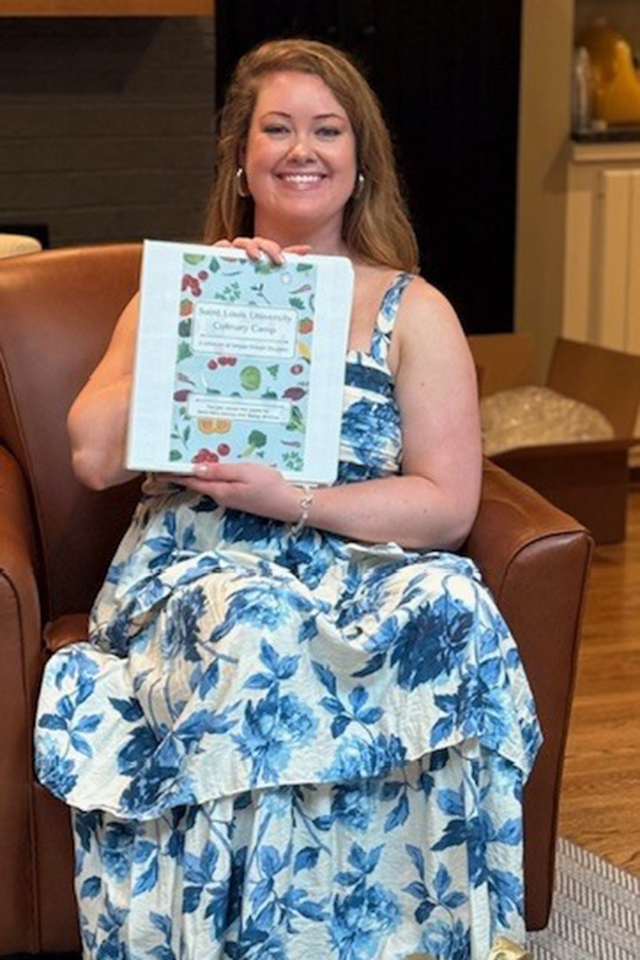 Mary Kate Keenoy with her recipe book A woman in a blue-patterned dress sits in a chair. She holds a recipe book and smiles for a photo indoors.