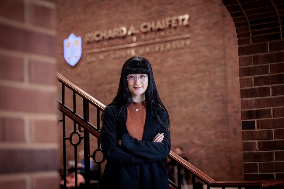 Woman with arms crossed, long, black hair and wearing glasses poses for a photo in front of a railing. 