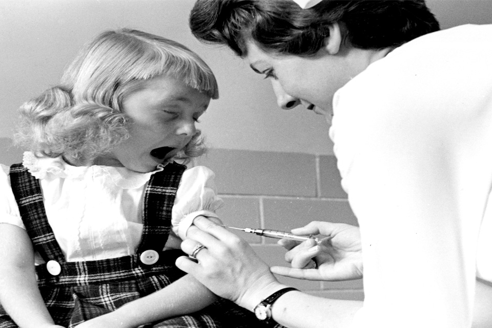 In this black and white vintage image, a young girl makes a face as a nurse injects her with a shot.