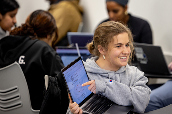 An undergraduate student holds a laptop and points to a document displayed on the laptop screen while smiling at another student.