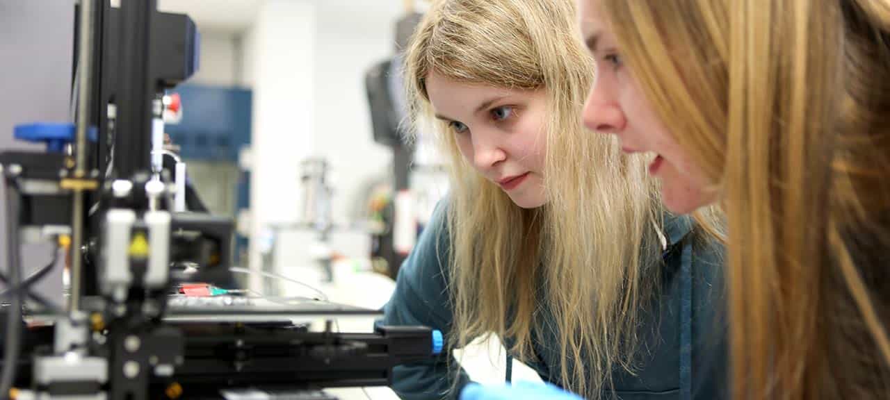 Two female students with long blonde hair lean in close to study a piece of equipment in a laboratory.